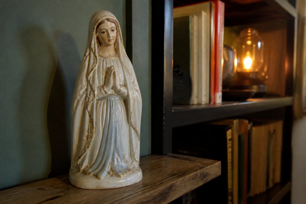 Close-up van een gipsen Mariabeeld uit de jaren 1920-1930 met zachte gezichtstrekken, elegant geplaatst op een houten plank naast een moderne boekenkast met warme lampverlichting