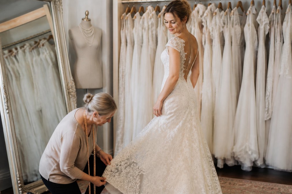 Seamstress adjusting the hem of a lace gown as a bride tries on one of many vintage wedding dresses in a boutique.