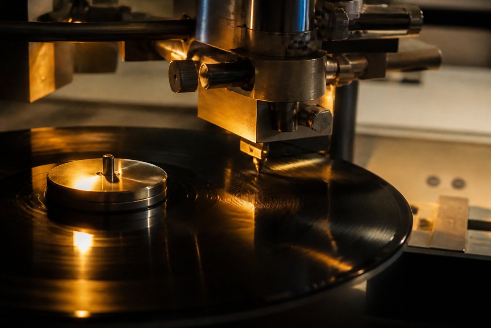 Close-up of a vinyl cutting lathe engraving a lacquer master in a dim professional mastering studio, engineer's hand adjusting a monitoring fader, warm amber lighting on the metal cutting head
