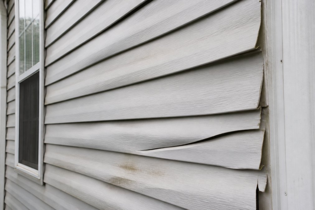 Close-up of warped and buckled vinyl siding on a home exterior, showing visible heat damage and panels pulling away from the wall