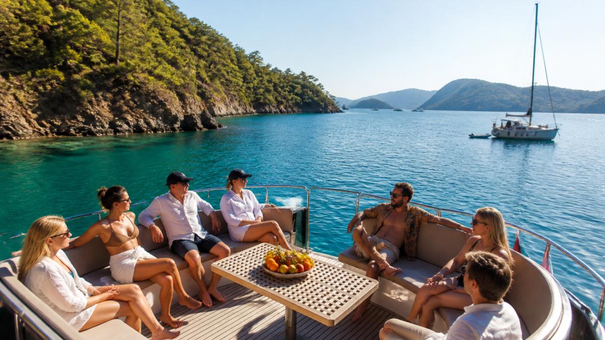 Turist grubu tekne güvertesinde oturmakta, turkuaz Akdeniz denizi ve uzakta yeşil koylar görülmekte, sabah güneş ışığında huzurlu deniz manzarası