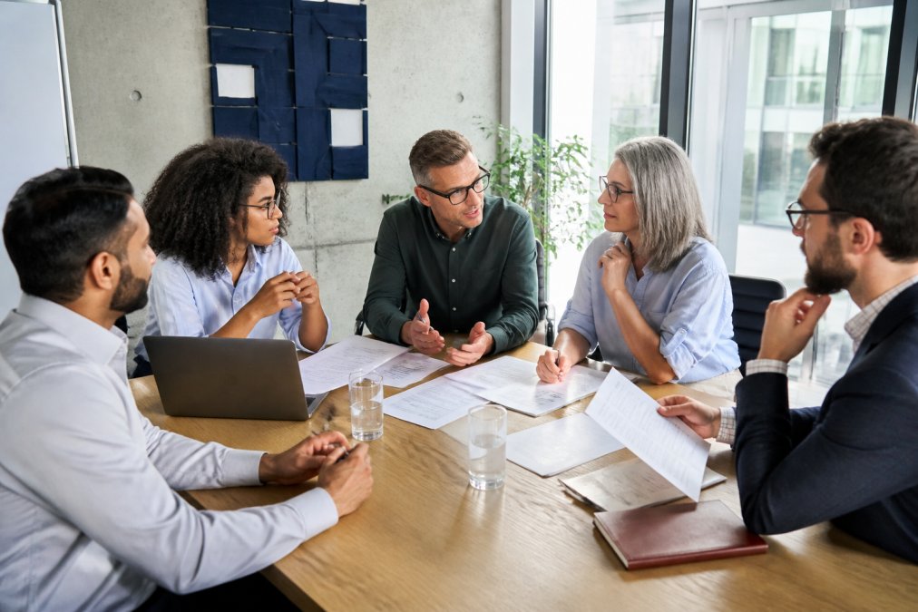 Business leaders and managers in a conference room reviewing training materials and discussing strategy around a table with laptops and documents, focused on planning