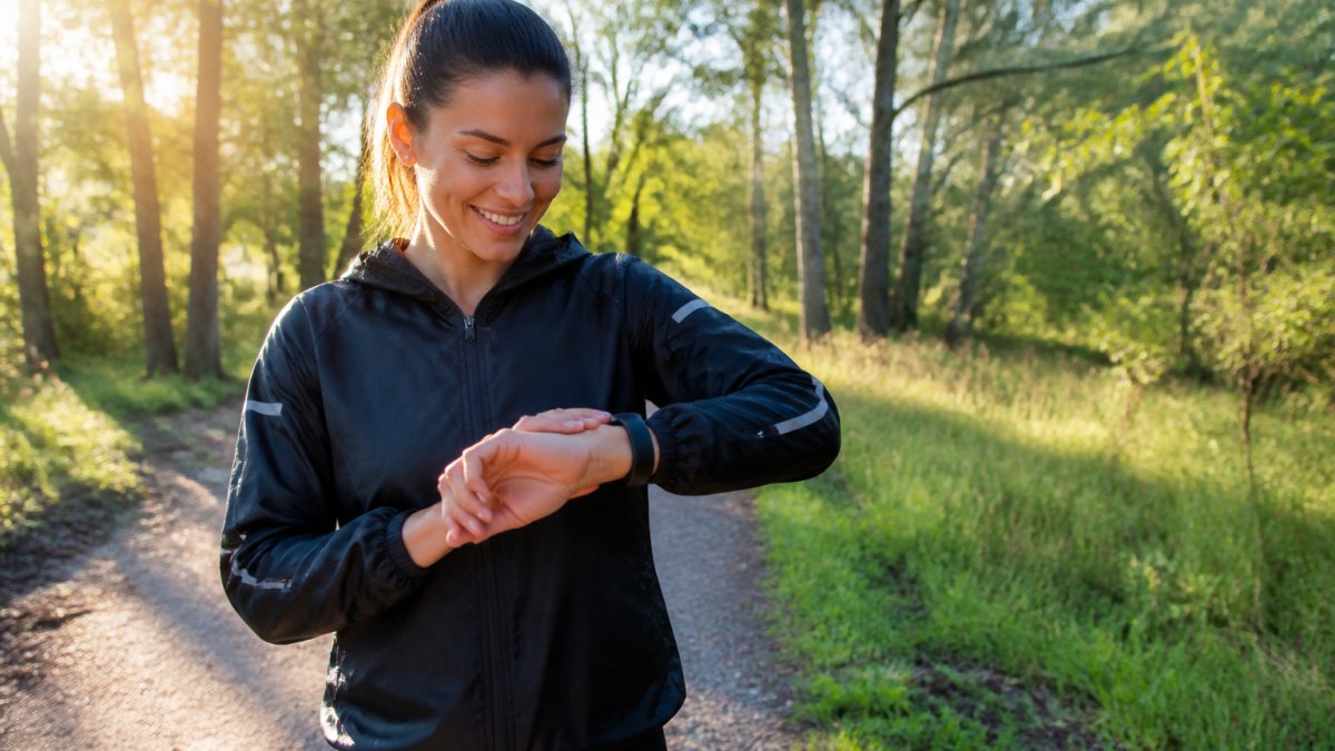 Runner outdoors on a sunny morning wearing trainers and fitness gear, smiling whilst checking their running watch to log distance on their smartphone