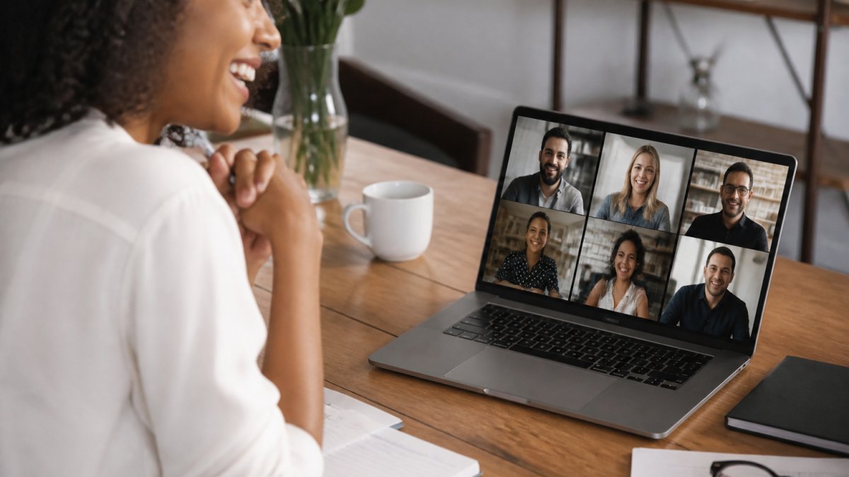 Diverse sales team members smiling and engaged during a virtual meeting, visible on laptop screens with coffee cups and notebooks on their desks, natural office lighting
