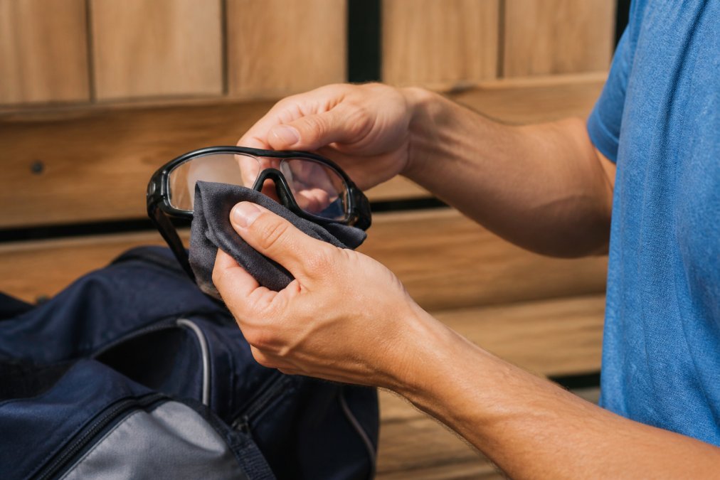 Close-up of hands cleaning prescription sports goggles with microfiber cloth in outdoor locker room setting, proper maintenance technique visible with soft cloth material and frame detail