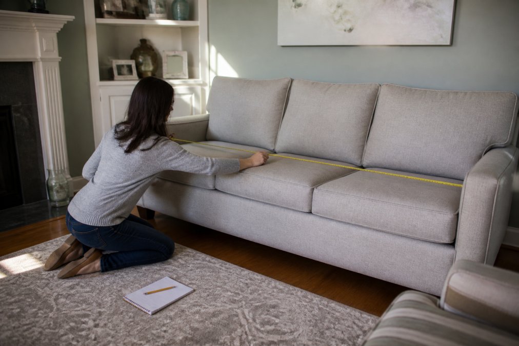A person kneeling on a hardwood living room floor with a tape measure extended across a sofa, notebook and pencil beside them, measuring wall width in soft afternoon light
