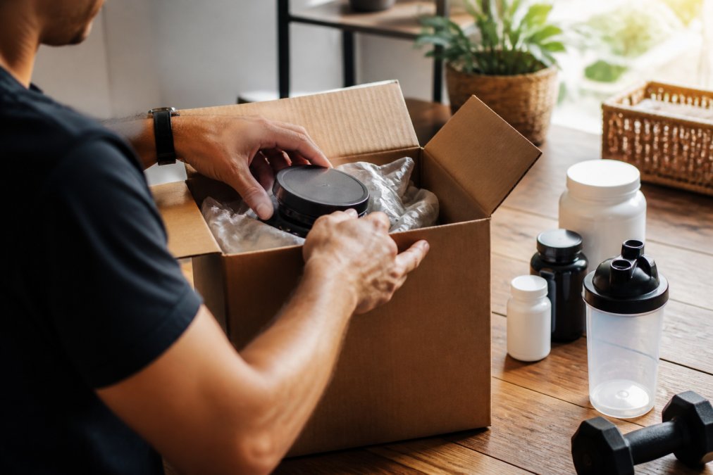 Close-up of hands opening a supplement package at a home workspace, with a laptop and wellness products visible nearby, natural afternoon lighting