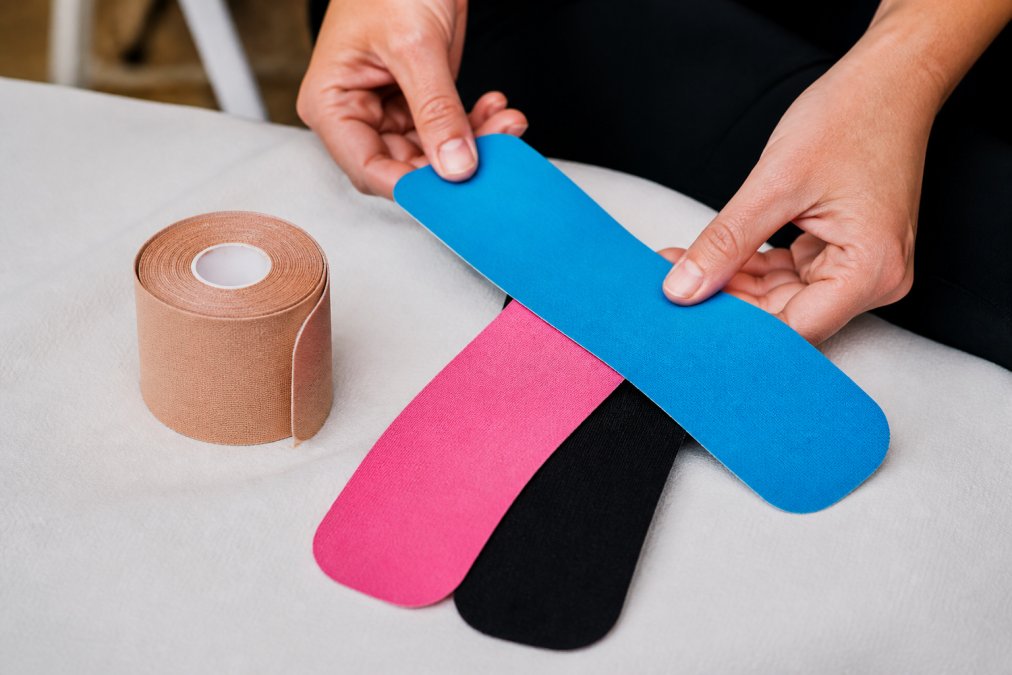 Close-up of hands showing kinesiology tape strips in different colors (blue, pink, black) arranged on a white medical surface next to a roll of elastic tape