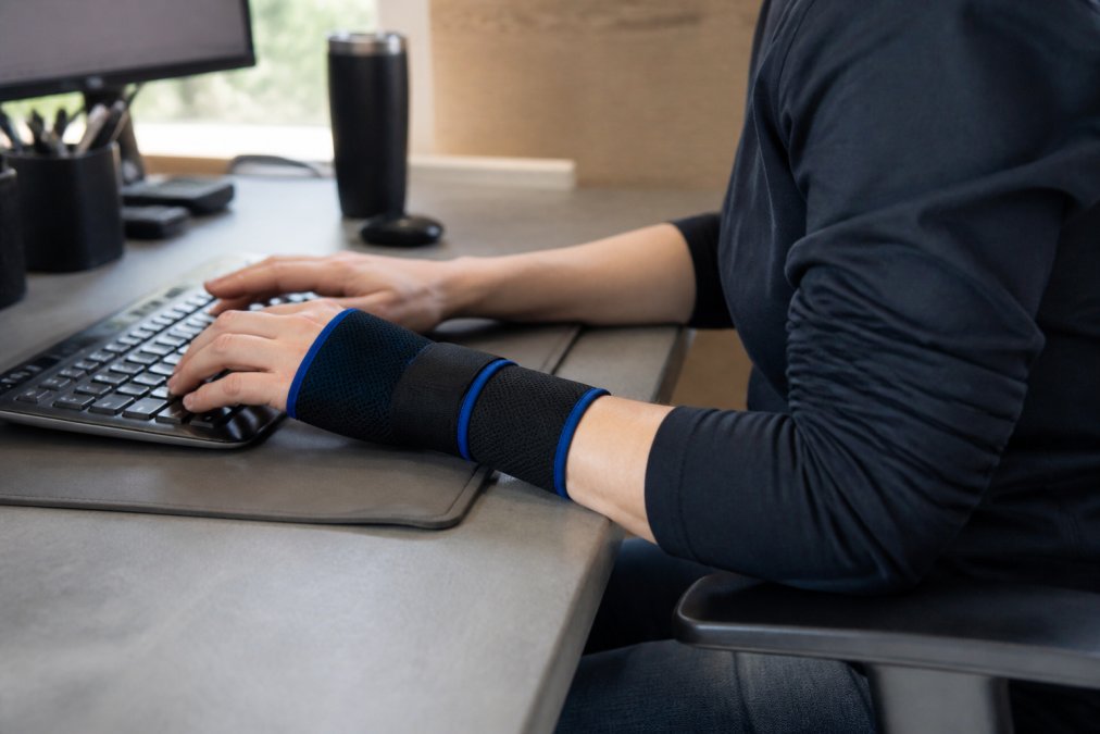 Person wearing a cold compression sleeve on their wrist while sitting at an ergonomic desk, typing on a keyboard, with the sleeve clearly visible showing proper fit and positioning during daily work activities