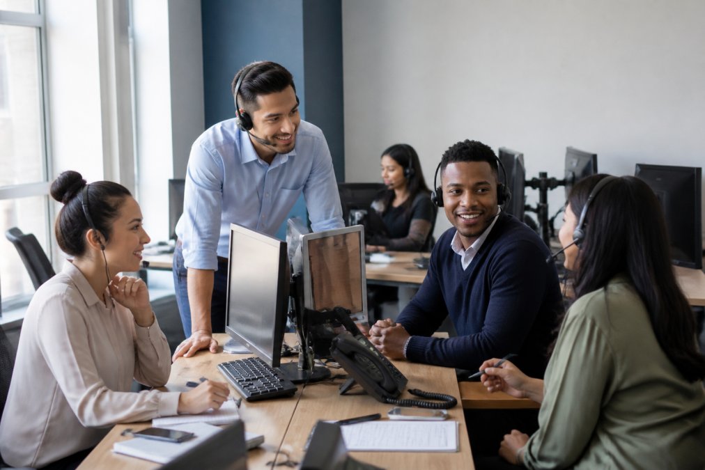 Sales team in modern office collaborating around a desk with multiple monitors displaying customer data and call information, professional headsets visible, natural office lighting