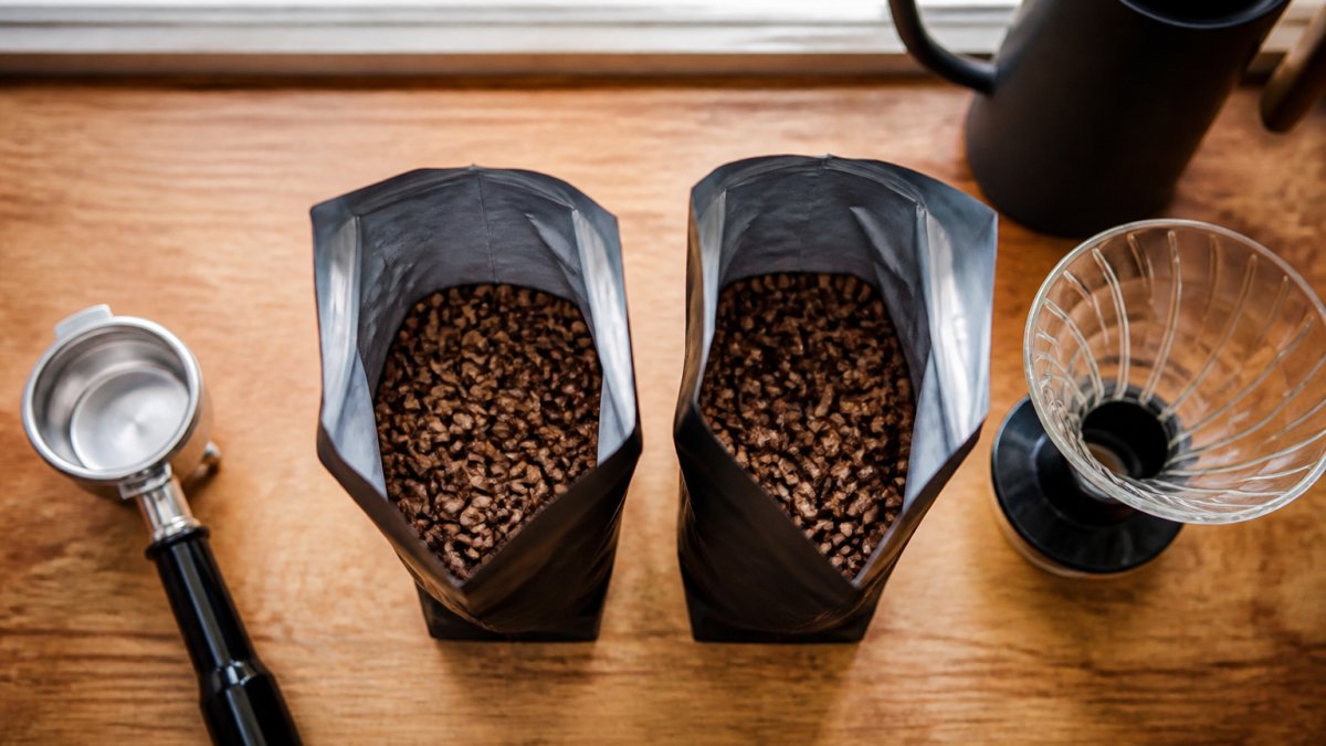 Overhead shot of two open bags of coffee beans side by side on a wooden kitchen counter, with a portafilter and a pour-over dripper nearby, natural window light