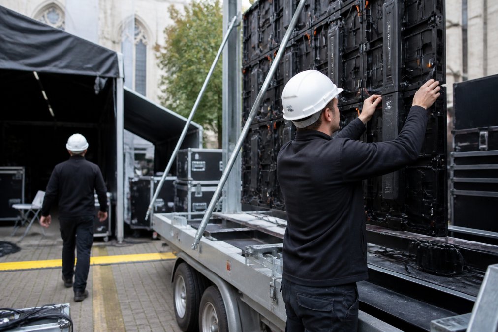 Technische medewerkers in veiligheidshelmen die een mobiel LED-scherm op aanhangwagen opzetten, stroomkabels aansluiten in regieruimte, professionele installatie in volle gang op evenementlocatie in Antwerpen