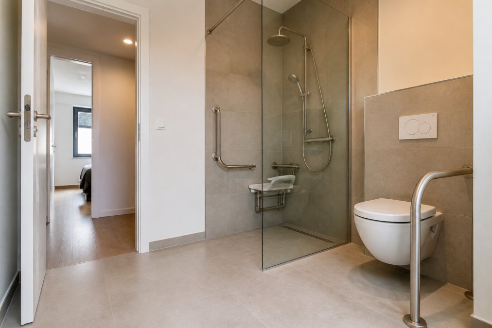Een moderne, lichte badkamer met inloopdouche zonder drempel, grijpstangen in neutrale kleuren, een comfortabel toilet, en brede deuren die zichtbaar zijn, alles ontworpen voor veiligheid en gemak