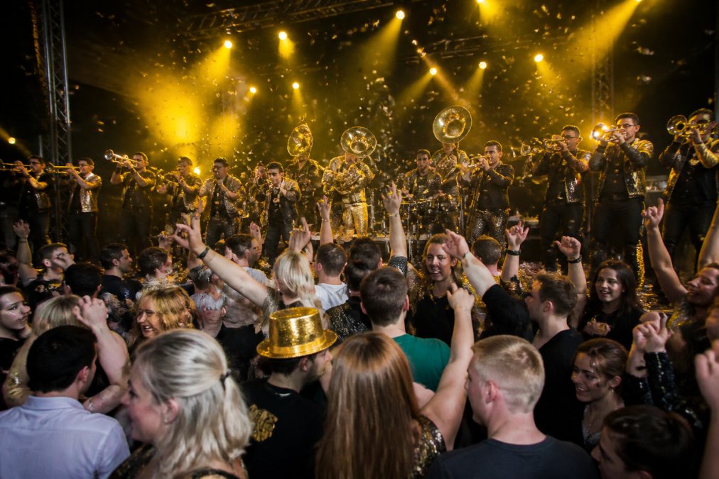 Energieke brassband op een groot podium met blazers en drummers in volledige actie, publiek danst en springt in de voorgrond onder warm geel podiumlicht, confetti in de lucht