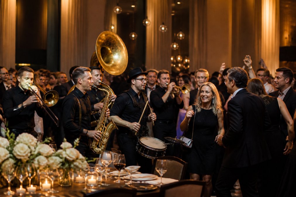 Professioneel geklede muzikanten van een brassband op een bedrijfsfeest, interacterend met geklede gasten die dansen en glimlachen rond een elegant ingericht diner-podium, zachte gouden verlichting