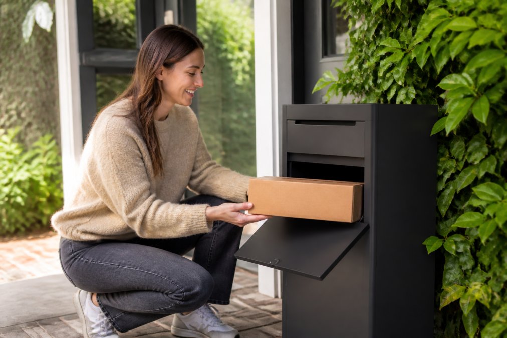 Iemand die thuis werkt ontvangt een pakket via een moderne pakketbrievenbus aan de voordeur, met een glimlach van tevredenheid