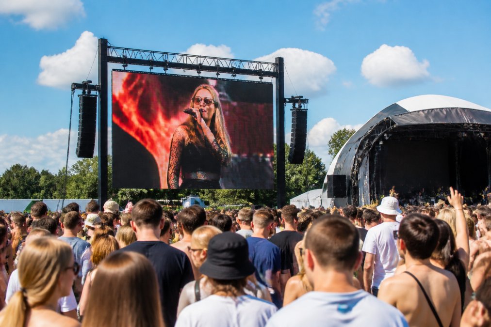 Groot professioneel LED-scherm buiten op druk openluchtfestival in fel zonlicht, met heldere, levendige beelden en duidelijk zichtbare tekst ondanks sterke daglichtverhoudingen, omringd door bezoekers