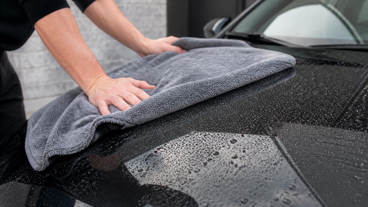 Close-up van iemand die een auto met een grote microvezel droogdoek voorzichtig droogt in de schaduw, waterdruppels glinsteren op de lak
