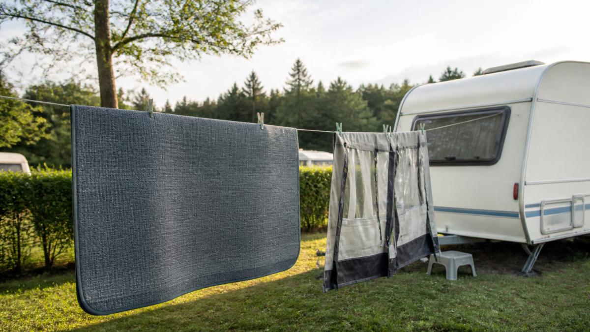 Ein Vorzeltteppich hängt zum Trocknen über einer Wäscheleine auf einem Campingplatz, daneben steht ein Wohnwagen, die Windschürze ist abgenommen und seitlich aufgehängt, weiches Nachmittagslicht