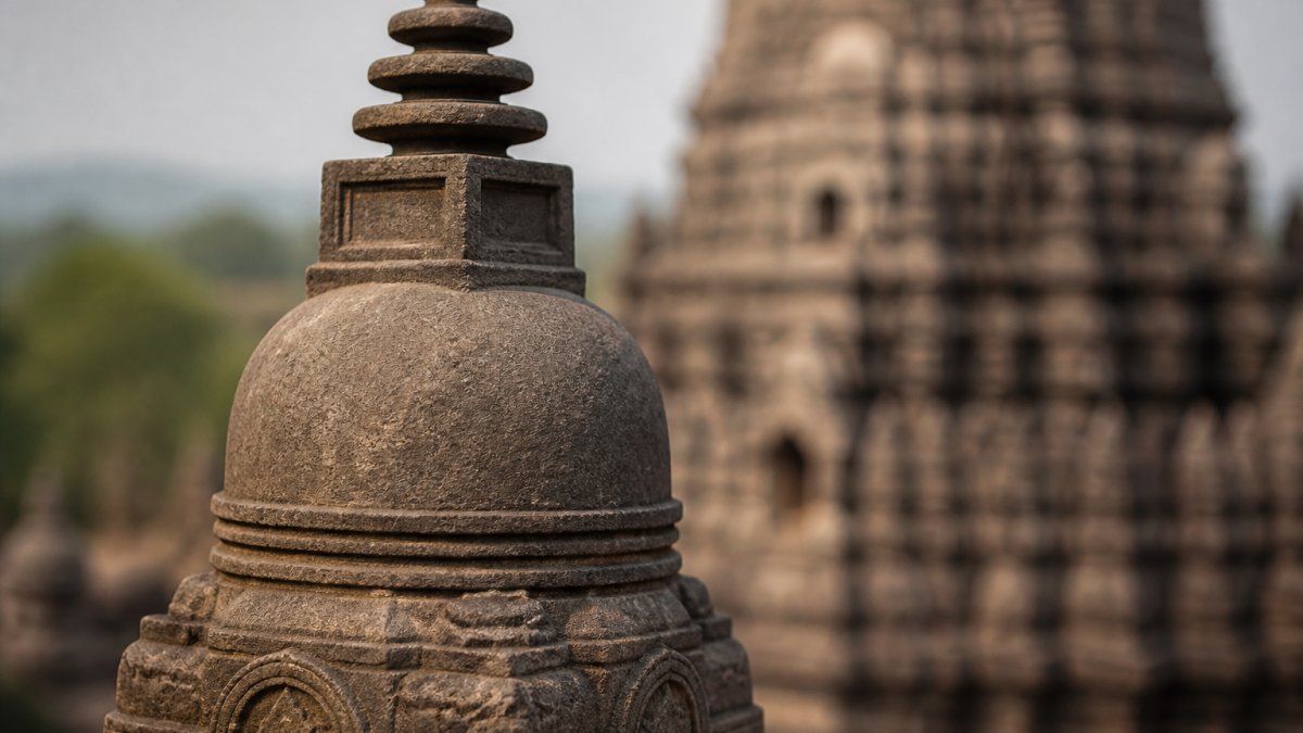 Close-up of a small carved stone votive stupa showing the dome, square harmika, and stacked chattra discs, with a larger monument blurred in the background