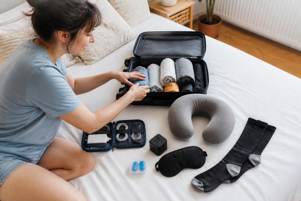 Flat lay photograph of travel accessories neatly arranged on a white bed: a navy cable organizer, black universal adapter, gray memory foam neck pillow, black 3D sleep mask, blue silicone earplugs in case, and black compression socks, all beside a compact carry-on bag in bright natural daylight