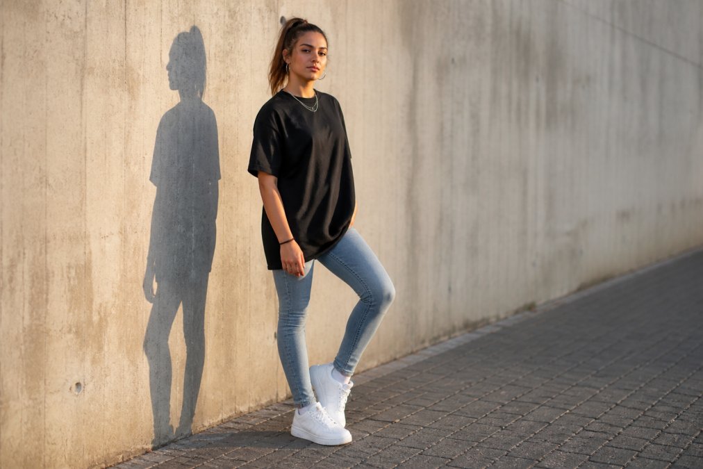 A young woman wears an oversized black streetwear T-shirt paired with fitted blue jeans and white sneakers. She stands confidently against a concrete wall in an urban setting, with the late-afternoon light casting a long shadow. The photo emphasizes how the loose top still creates a flattering silhouette.
