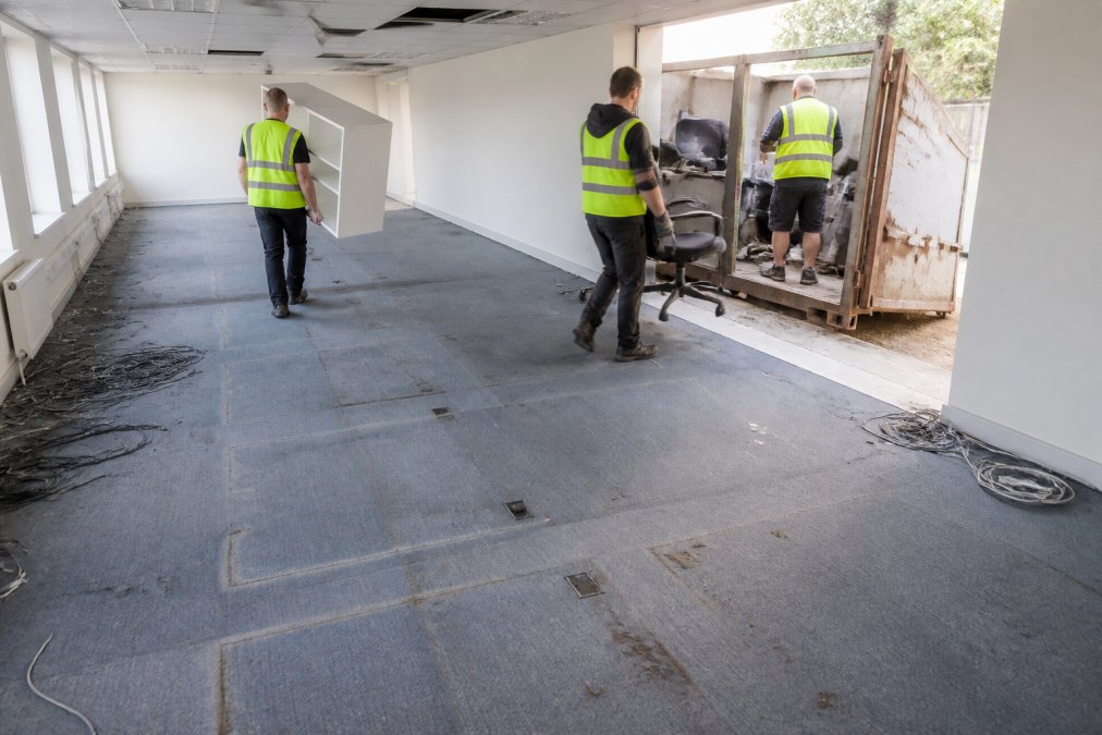 Pracovníci v reflexních vestách vynášejí kancelářské židle a skříně z prázdné kancelářské budovy do přistaveného kontejneru, na podlaze jsou vidět značky po nábytku a zbytky kabeláže