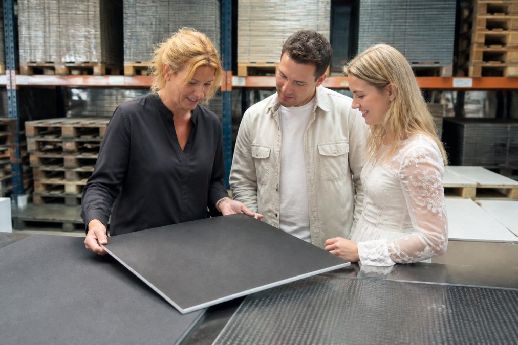 Petra Keizer van Discovloer Huren bekijkt samen met een bruidspaar de modulaire panelen van een duurzame dansvloer in een lichte showroom, met op de achtergrond stapels gerecyclede panelen