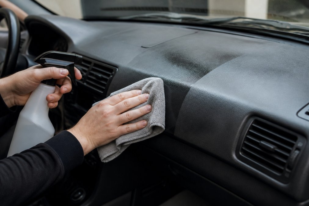 Close-up van iemand die met een microvezeldoek en een sprayfles het kunststof dashboard van een auto reinigt, met zichtbaar verschil tussen een dof en een glanzend gedeelte