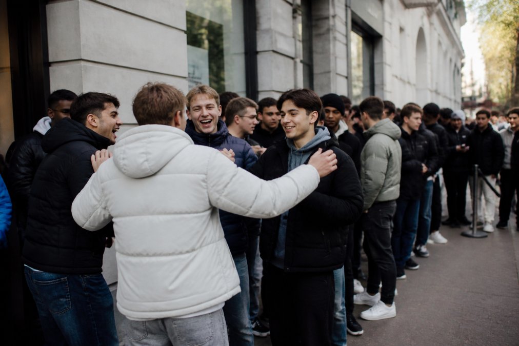 Groep jongvolwassenen staat buiten een winkel te wachten voor een streetwear drop, telefoons in hand, energieke sfeer met anticipatie en opwinding terwijl ze elkaar aanstoten
