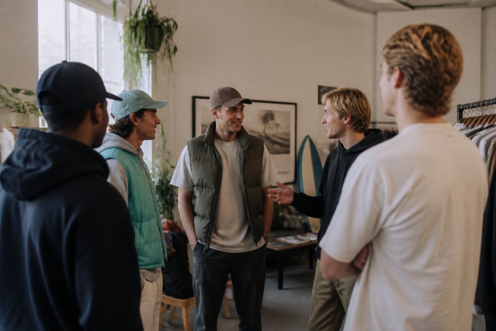 Een groep jongvolwassenen in unieke, kleurrijke streetwear-outfits die samen in een moderne, creatieve ruimte staan met graffiti-achtige wandschilderingen, duidelijk in gesprek en lachend, verbonden door hun gezamenlijke passie voor mode