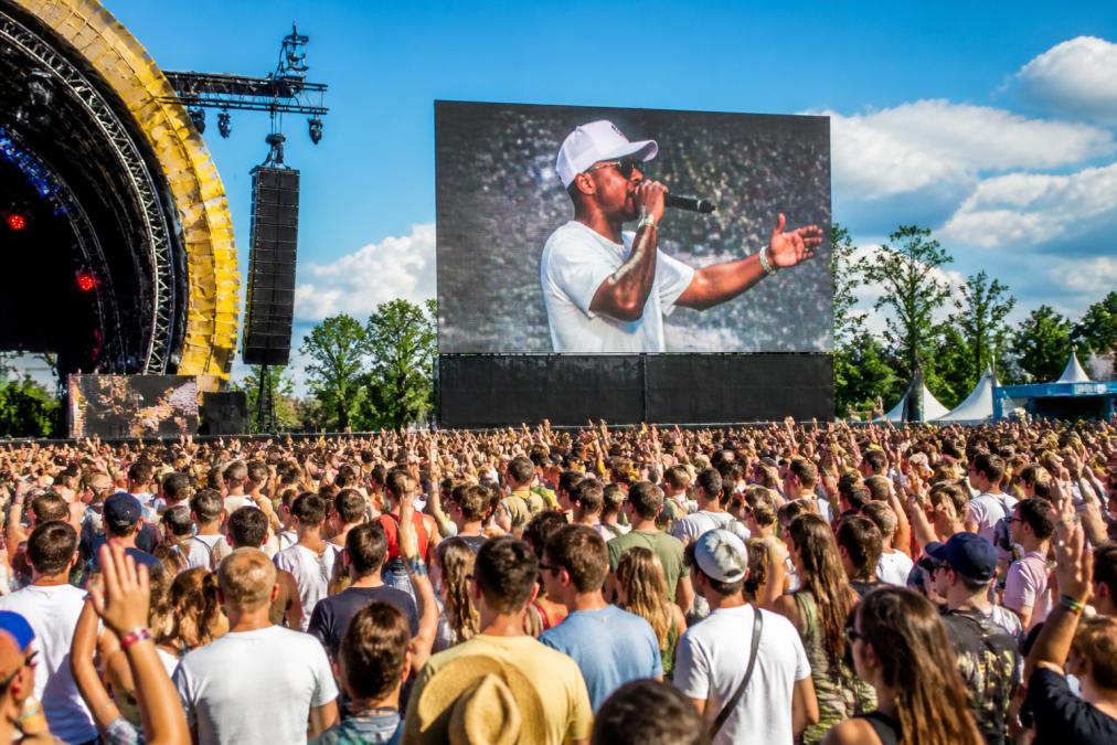 Groot LED-scherm op een druk festivalterrein met honderden bezoekers die naar het scherm kijken, met levendige kleuren en helder beeld overdag, veel beweging en energie
