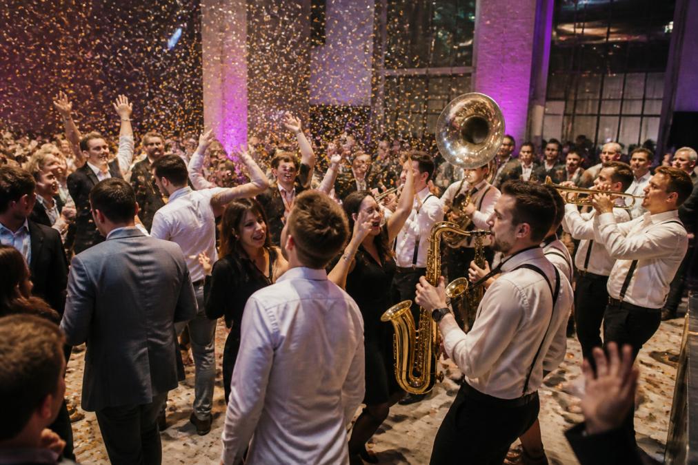 Energieke brassband in volle actie op bedrijfsfeest met muzikanten die het publiek aanzetten tot dansen, confetti in de lucht, en gasten die enthousiast op de dansvloer bewegen in feestelijke sfeer