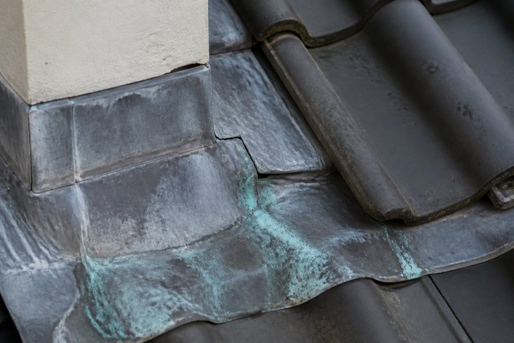 Close-up van beschadigd loodwerk op dakrand met zichtbare groene corrosielaag, scheurvorming en verkleuringen tegen grijze dakpannen