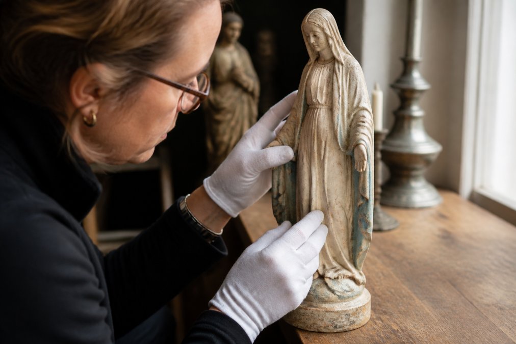 Handen van een expert die voorzichtig de details van een antiek gipsen Mariabeeld onderzoeken, met focus op de signatuur, patina en handgesneden elementen in natuurlijk licht