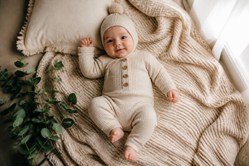 Baby wearing a cream waffle romper and matching knit hat lying on a neutral blanket in natural window light