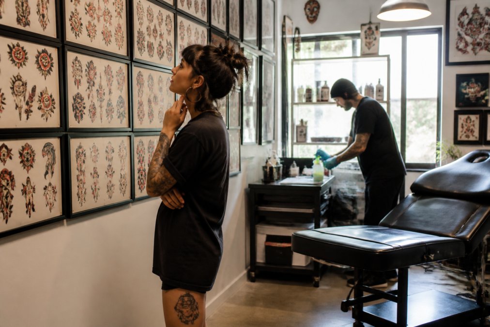 A bright, modern tattoo studio interior with a client reviewing flash art on a wall while a tattoo artist prepares a station in the background