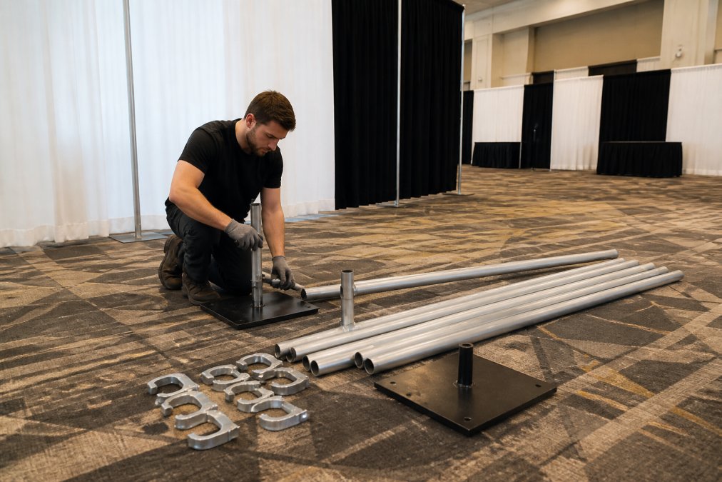 Professional installer assembling pipe and drape hardware components on event venue floor, showing uprights, crossbars, base plates, and C-clamps positioned and ready for connection, with natural overhead lighting