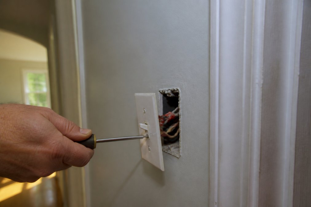 Homeowner using a screwdriver to remove a warm light switch plate to inspect the electrical wiring inside.