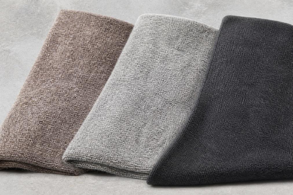 Close-up of three different thermal base layer fabric swatches (alpaca, merino, and synthetic blend) laid flat on a neutral surface, showing distinct fiber texture and density differences under natural light