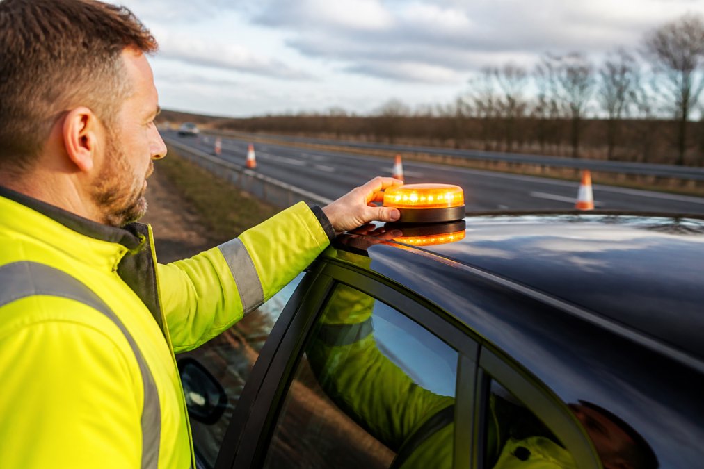Warnlight vs LED-GPS: Safety Comparison 2026 Close-up of a magnetic emergency beacon being attached to the roof of a vehicle parked on a motorway hard shoulder, with the beacon's amber light visible flashing in daylight