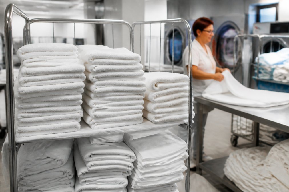 Gestapeld schoon wit linnengoed en handdoeken op een kar in de wasserij van een hotel of zorginstelling, met een medewerker op de achtergrond die textiel vouwt