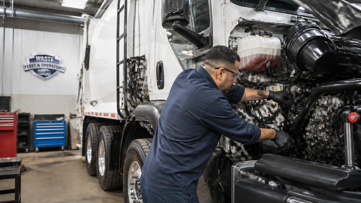 Professional technician performing hands-on inspection of waste collection truck, examining engine compartment and hydraulic systems in a maintenance bay with bright overhead lighting