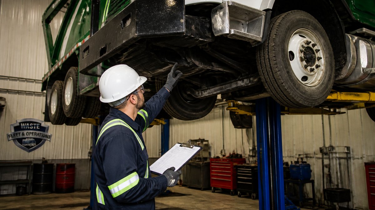 A fleet maintenance technician in safety gear inspecting the undercarriage of a waste collection truck in a maintenance bay, clipboard in hand, with the truck raised on a lift