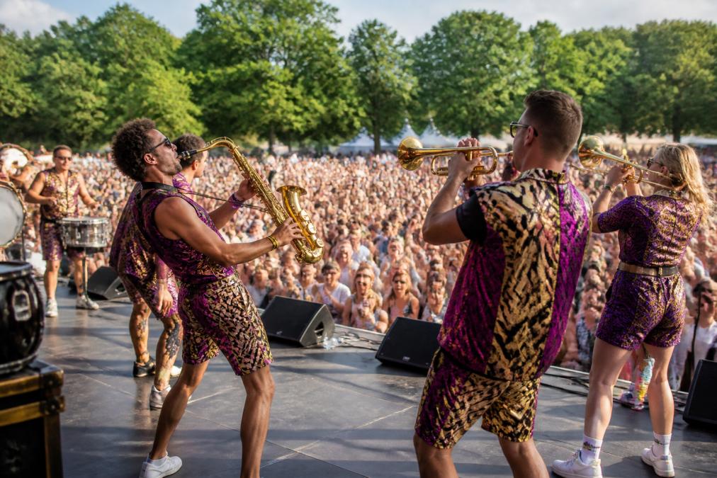 Een energieke brassband met blazers en slagwerk die optreedt op een podium voor een groot publiek, met kleurrijke kleding en zichtbare interactie met de menigte