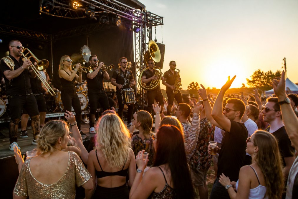 Energieke brassband in actie op een festivalpodium, koperblazers en slagwerkers in beweging, publiek dat danst en juicht op de voorgrond, warme avondzon