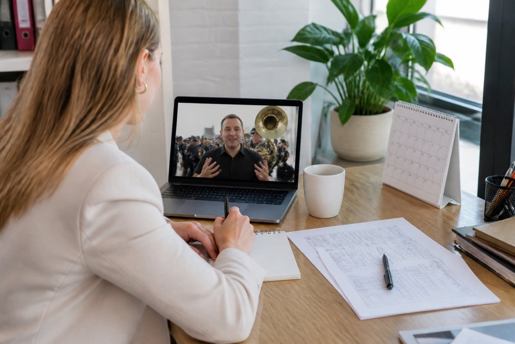 Eventplanner zittend aan bureau met laptop en offerte-documenten, in videocall met brassband-manager, notitieblok en kalender zichtbaar op bureau