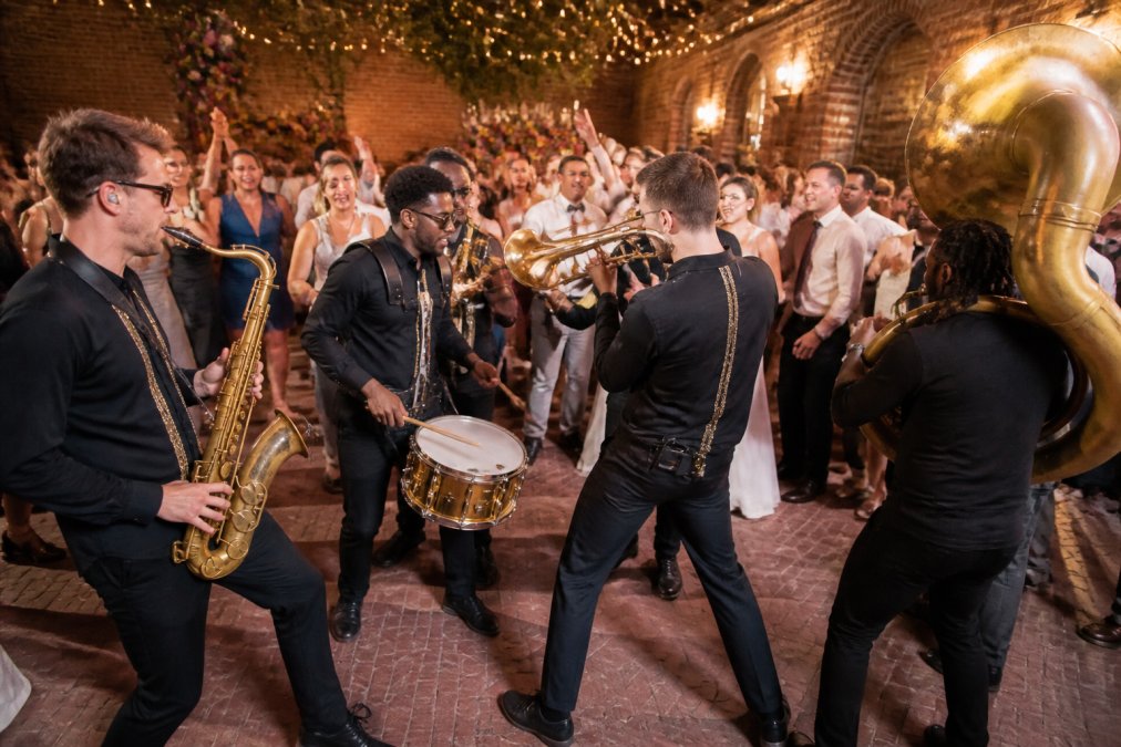 Een energieke brassband met blazers en percussie die optreedt op een feestelijk versierde trouwlocatie, gasten dansen en klappen mee op de achtergrond