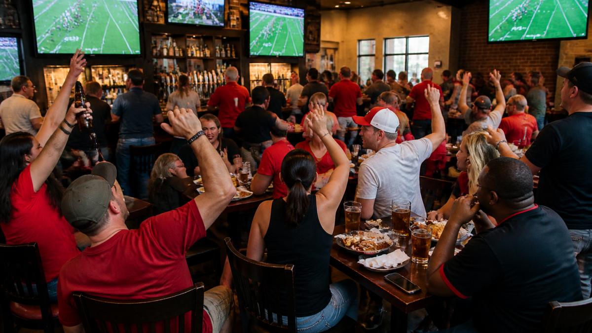 Lively sports bar packed with college football fans watching multiple large screens, people seated at tables and standing by the bar, holding beers and appetizers, cheering during a game, with warm lighting and energy