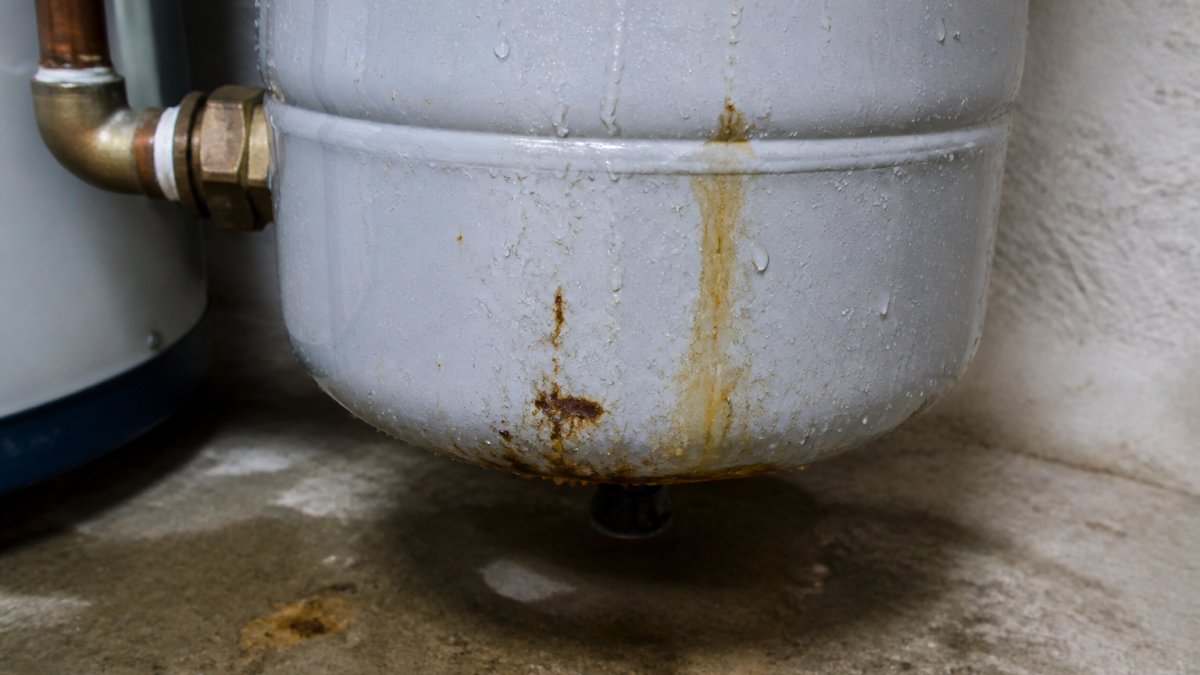 Close-up of water droplets and rust stains on a metal expansion tank, showing visible corrosion and wet spots on the tank surface and surrounding floor